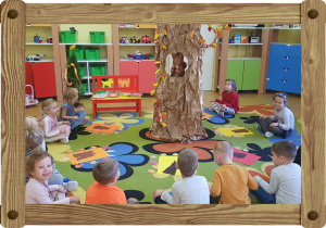 Dzieci siedzą na dywanie. W środku stoi duże jesienne drzewo.